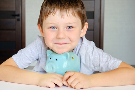 a cute blue-eyed boy with a smile on his face hugs his piggy bank, a blue pig. The child looks at the camera, horizontal photoの写真素材