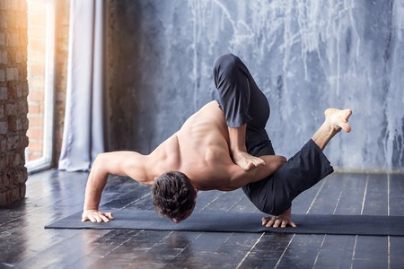 Young man doing an asana ashtavakrasana. Hand standing pose. Yogi master workout on black mat on...