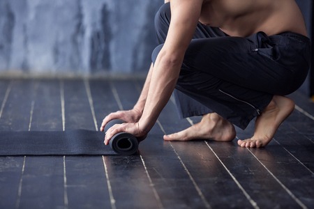 Young yogi men rolling mat after a yoga on black wooden floorの写真素材