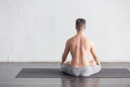 A young strong man doing yoga exercises - lotus pose and meditates with padmasana legs. Studio full length shot over white brick background and black floor, copy space, back view.の写真素材