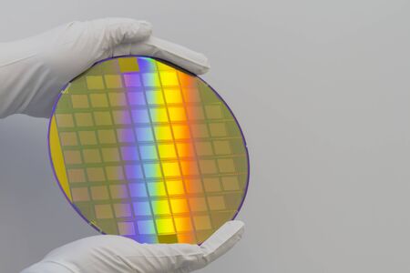 Hands in white gloves holding a silicon wafer with integrated circuits on a white background.
