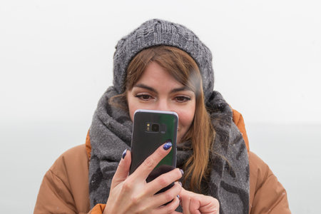 A portrait of a girl in a winter hat and a winter jacket with a scarf holds a smartphone and takes a photo.の写真素材