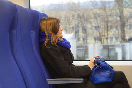 A young brunette woman sits and looks out the train window at the cityscapes. Holding a blue handbag. A blue scarf and blue seats. A smile on his face.の写真素材