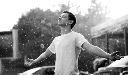 teenage boy standing under rain in the city anjoying rain and smiling. Black and white image. There is an street behind himの写真素材