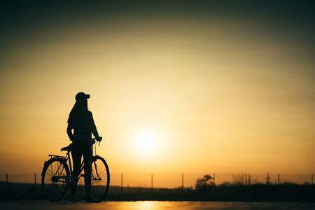 beautiful hipster girl with a bicycle on the road during sunsetの写真素材