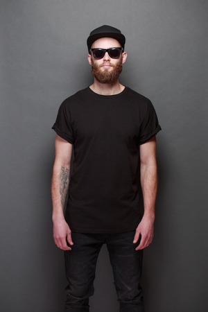 Hipster handsome male model with beard wearing black blank t-shirt with space for your design over gray backgroundの写真素材