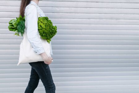 Woman holding an eco bag filled with grocery. Vegetables and fruits are hanging from a bag. Ecology or environment protection concept. White eco bag for mock up.の写真素材