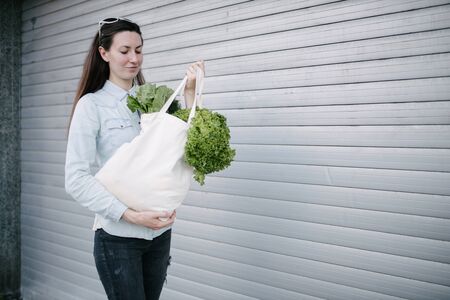 Woman holding an eco bag filled with grocery. Vegetables and fruits are hanging from a bag. Ecology or environment protection concept. White eco bag for mock up.の写真素材