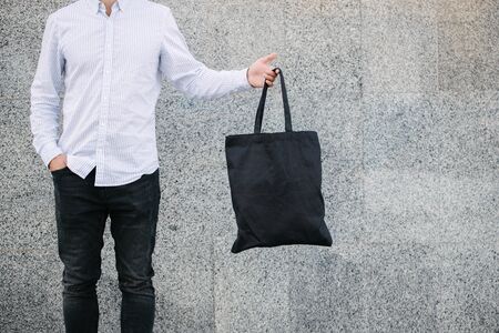 Young man holding black textile eco bag against urban city background. . Ecology or environment protection concept. Black eco bag for mock upの写真素材