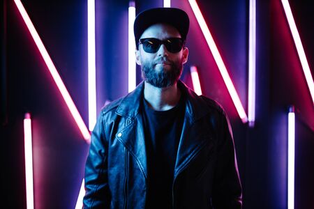 Hipster handsome man on the city streets being illuminated by neon signs. He is wearing leather biker jacket or asymmetric zip jacket with black cap, jeans and sunglasses.の写真素材