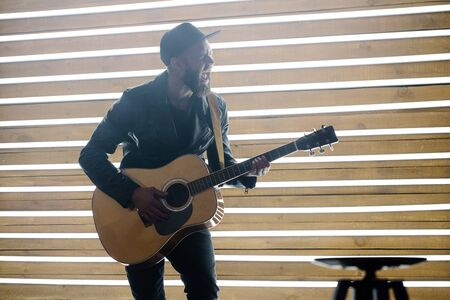Singer and guitar player singing on a stage with neon lights. He is a rocker and he is wearing leather biker jacket or asymmetric zip jacket with black cap, jeans.の写真素材