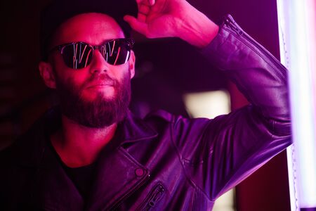 Hipster handsome man on the city streets being illuminated by neon signs. He is wearing leather biker jacket or asymmetric zip jacket with black cap, jeans and sunglasses.の写真素材