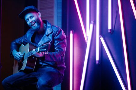 Singer and guitar player singing on a stage with neon lights. He is a rocker and he is wearing leather biker jacket or asymmetric zip jacket with black cap, jeans.の写真素材