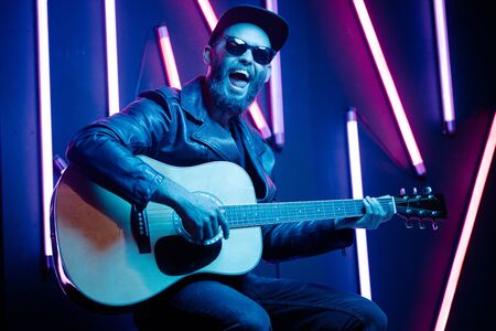 Singer and guitar player singing on a stage with neon lights. He is a rocker and he is wearing leather biker jacket or asymmetric zip jacket with black cap, jeans.の写真素材