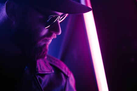 Hipster handsome man on the city streets being illuminated by neon signs. He is wearing leather biker jacket or asymmetric zip jacket with black cap, jeans and sunglasses.の写真素材