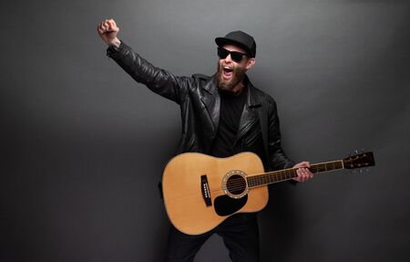 Singer and guitar player singing on a stage. He is a rocker and he is wearing leather biker jacket or asymmetric zip jacket with black cap, jeans.の写真素材