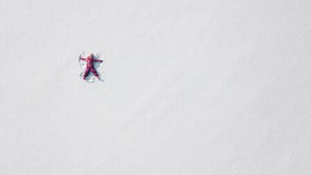 Kid on a snow making a snow angel. Aerial shot from above. She is wearing red clothesの写真素材