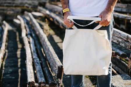 Young man holding white textile eco bag against urban city background. Ecology or environment protection concept. White eco bag for mock up.の写真素材