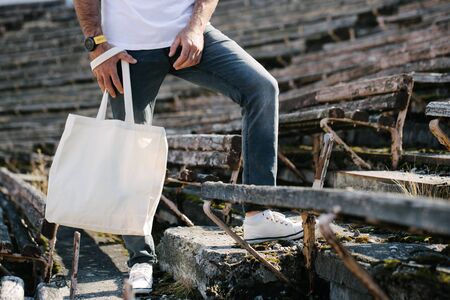 Young man holding white textile eco bag against urban city background. Ecology or environment protection concept. White eco bag for mock up.の写真素材