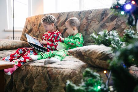 Children are reading a book next to a christmas tree wearing pajamasの写真素材