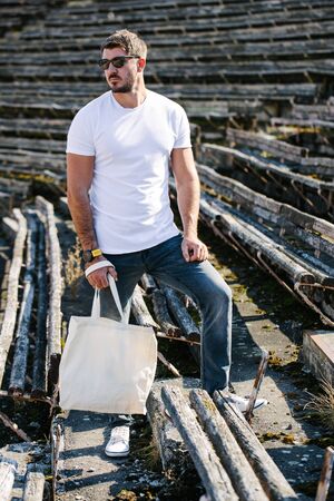 Young man holding white textile eco bag against urban city background. Ecology or environment protection concept. White eco bag for mock up.の写真素材