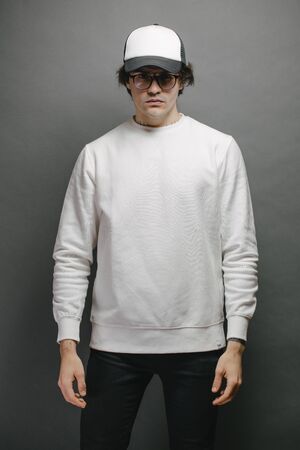 Man wearing blank white sweatshirt and empty baseball cap standing over gray background. Sweatshirt or hoodie for mock up, logo designs or design print with with free space.の写真素材