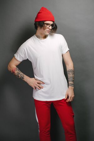 Hipster handsome male model with glasses wearing white blank t-shirt and red pants and a red hat with space for your logo or design over gray backgroundの写真素材