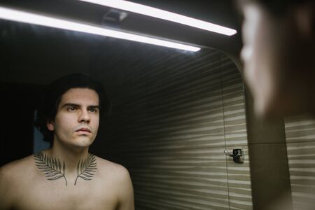 Young man in bathroom looking into a mirror at himself. He has a tattoo over his neckの写真素材