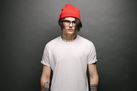 Hipster handsome male model with glasses wearing white blank t-shirt and red pants and a red hat with space for your logo or design over gray backgroundの写真素材