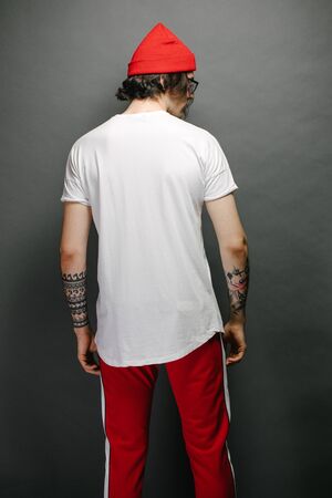 Hipster handsome male model with glasses wearing white blank t-shirt and red pants and a red hat with space for your logo or design over gray backgroundの写真素材