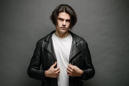 Man wearing leather biker jacket or asymmetric zip jacket with black cap, jeans and sunglasses. Handsome hipster man over gray background.の写真素材
