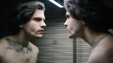 Young man in bathroom looking into a mirror at himself. He has a tattoo over his neckの写真素材