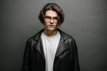 Man wearing leather biker jacket or asymmetric zip jacket with black cap, jeans and sunglasses. Handsome hipster man over gray background.の写真素材