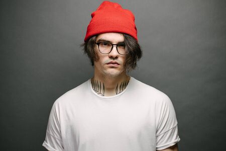Hipster handsome male model with glasses wearing white blank t-shirt and red pants and a red hat with space for your logo or design over gray backgroundの写真素材