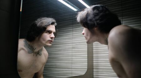 Young man in bathroom looking into a mirror at himself. He has a tattoo over his neckの写真素材