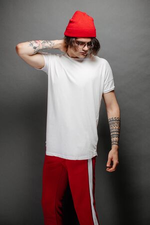 Hipster handsome male model with glasses wearing white blank t-shirt and red pants and a red hat with space for your logo or design over gray background.の写真素材