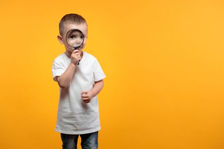 Boy looking into a magnifying glass and standing over yellow backgroundの写真素材