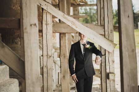 Fashion portrait of a groom outside. Half of his face is hidden behind a wooden plankの写真素材