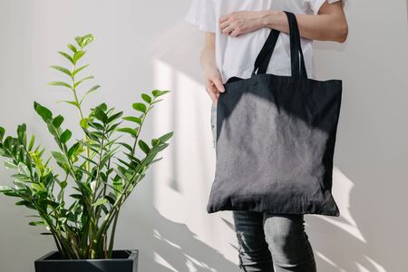 Young woman holding black textile eco bag against white wall. Ecology or environment protection concept. White eco bag for mock up.の写真素材