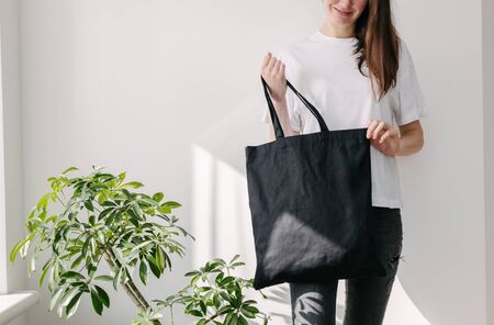 Young woman holding black textile eco bag against white wall. Ecology or environment protection concept. White eco bag for mock up.の写真素材
