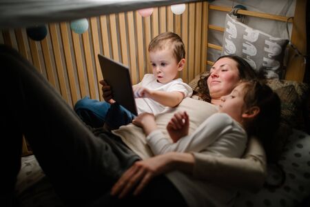 Children with mother watching a tablet pc and sitting in bed with garlands and having fun. Staying connected conceptの写真素材
