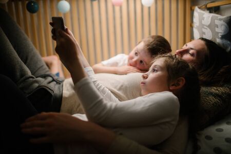 Children with mother watching a tablet pc and sitting in bed with garlands and having fun. Staying connected conceptの写真素材