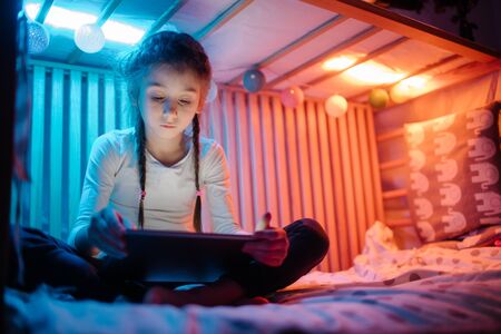 Child watching tablet pc at home. Girl watching a tablet and sitting in bed with garlands and having funの写真素材