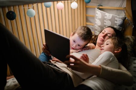 Children with mother watching a tablet pc and sitting in bed with garlands and having fun. Staying connected conceptの写真素材