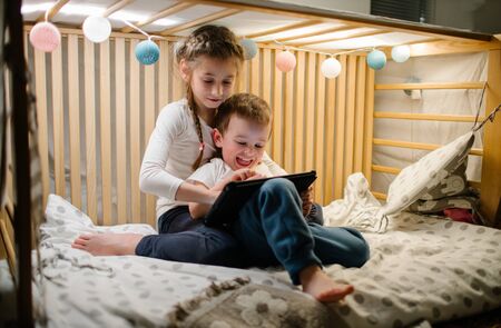 Children watching tablet pc at home. Brother and sister watching a tablet and sitting in bed with garlands and having funの写真素材
