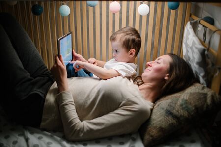 Children with mother watching a tablet pc and sitting in bed with garlands and having fun. Staying connected conceptの写真素材