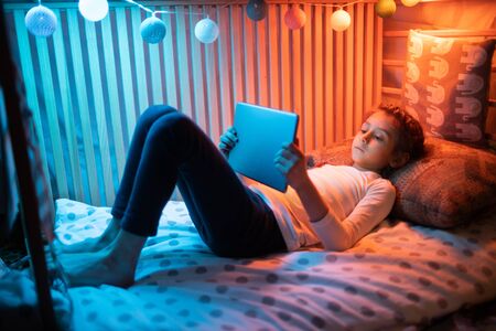 Child watching tablet pc at home. Girl watching a tablet and sitting in bed with garlands and having funの写真素材