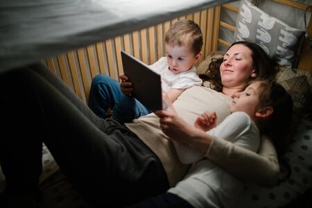 Children with mother watching a tablet pc and sitting in bed with garlands and having fun. Staying connected conceptの写真素材