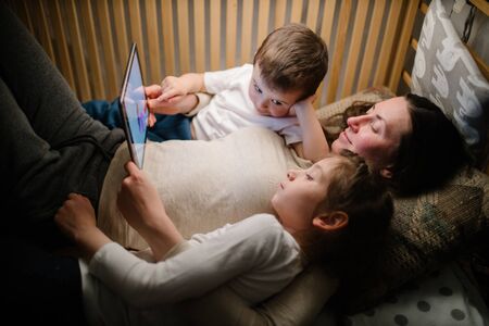 Children with mother watching a tablet pc and sitting in bed with garlands and having fun. Staying connected conceptの写真素材