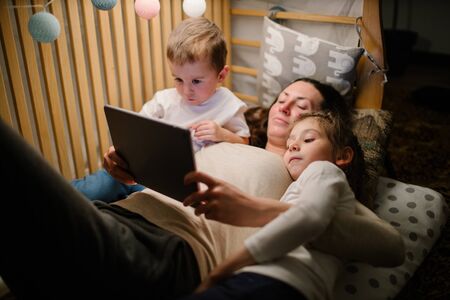 Children with mother watching a tablet pc and sitting in bed with garlands and having fun. Staying connected conceptの写真素材